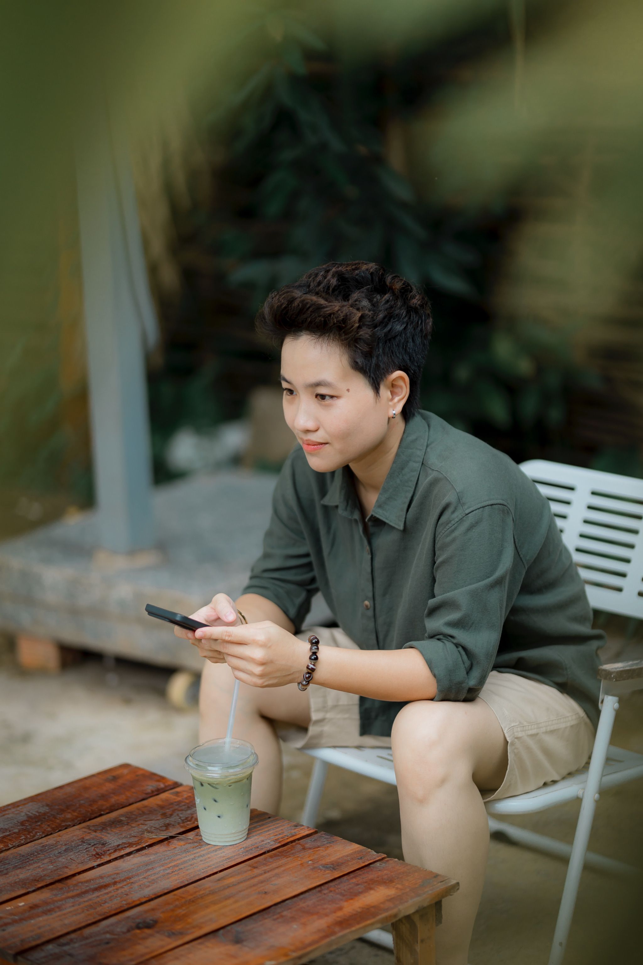 Young adult using smartphone outdoors, enjoying matcha drink in a relaxed setting.