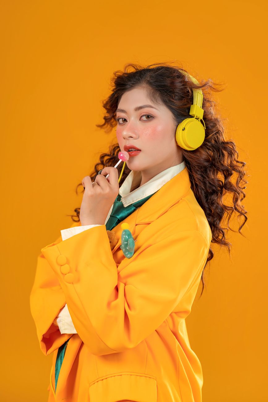 Fashionable woman in orange jacket and headphones holding lollipop.