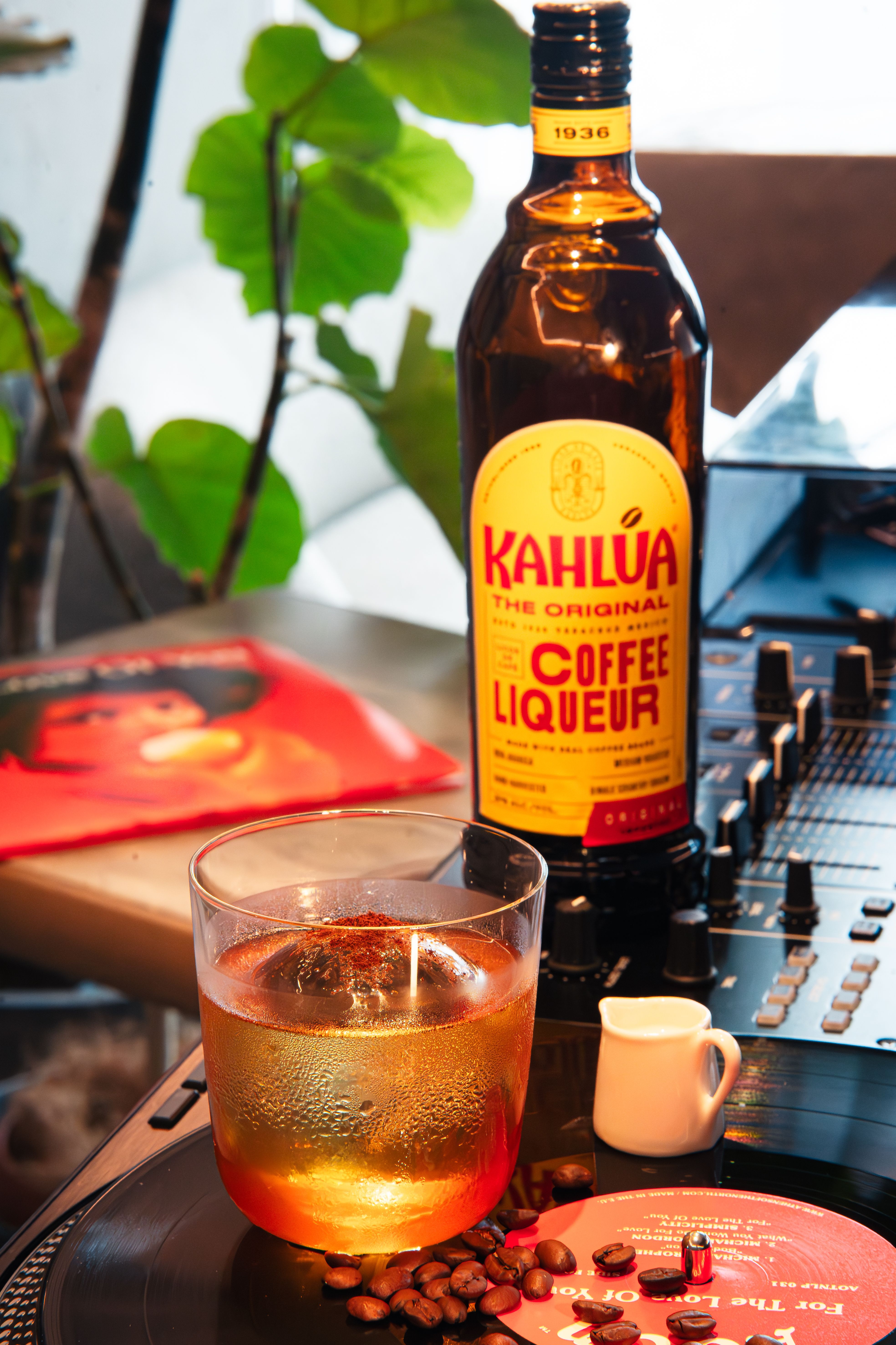 A bottle of Kahlua coffee liqueur next to a White Russian cocktail and scattered coffee beans on a dark wood surface.