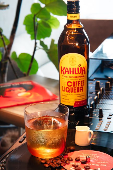 A bottle of Kahlua coffee liqueur next to a White Russian cocktail and scattered coffee beans on a dark wood surface.