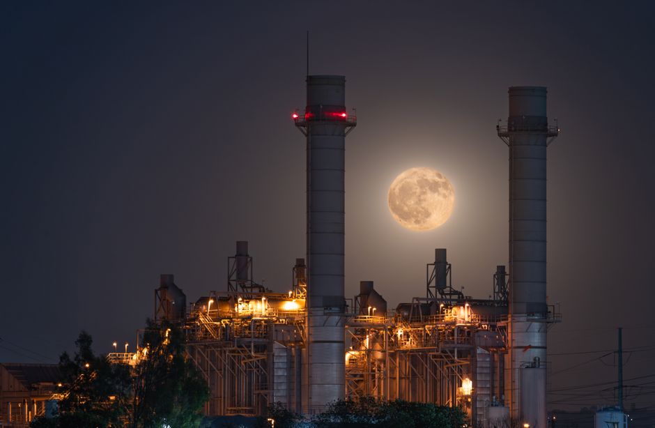 A dramatic full moon rising behind an illuminated industrial power plant at night.