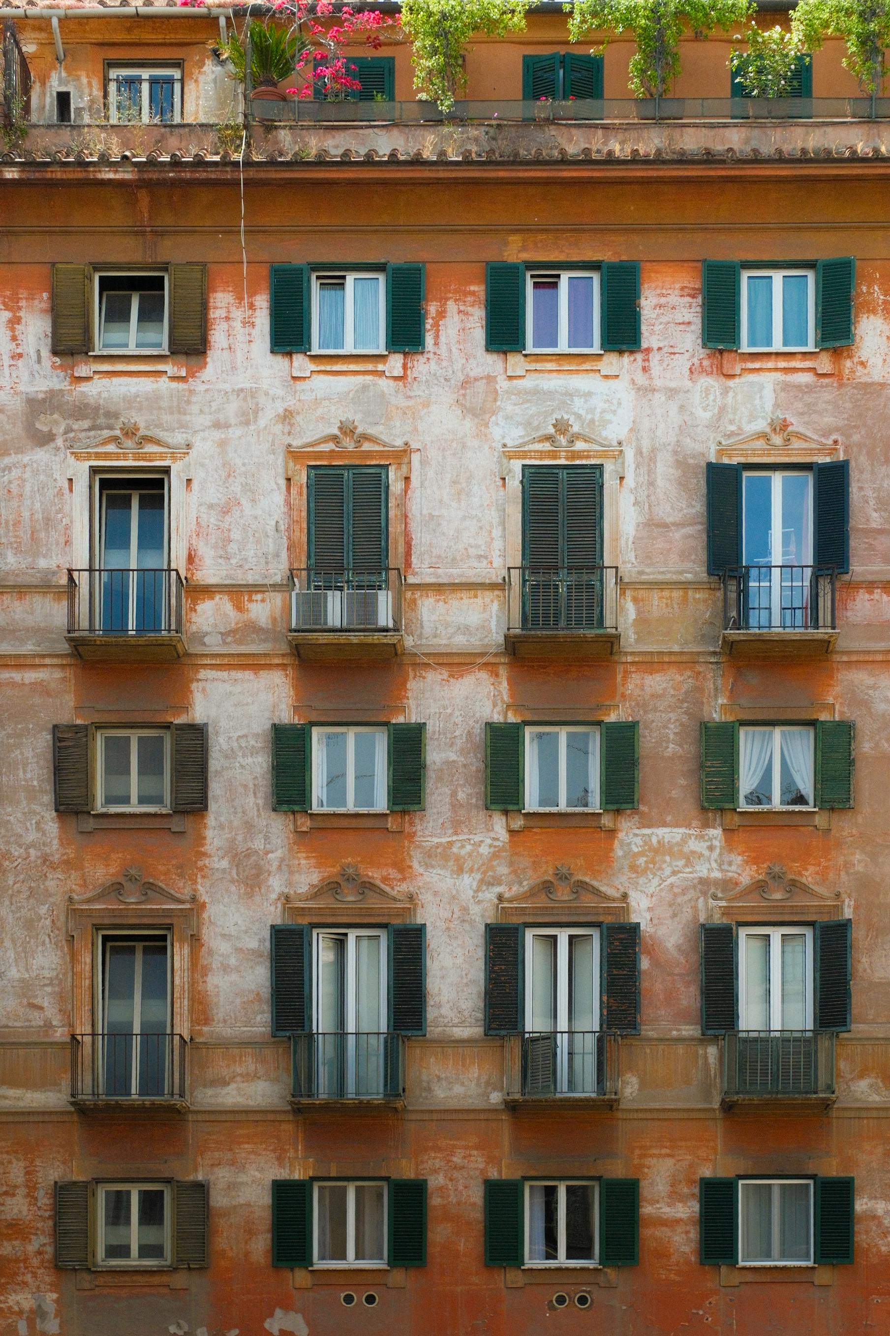 Gratis Affascinante facciata di un edificio usurato dal tempo, con persiane verdi e fiori dai colori vivaci, che catturano l'architettura classica. Foto a disposizione