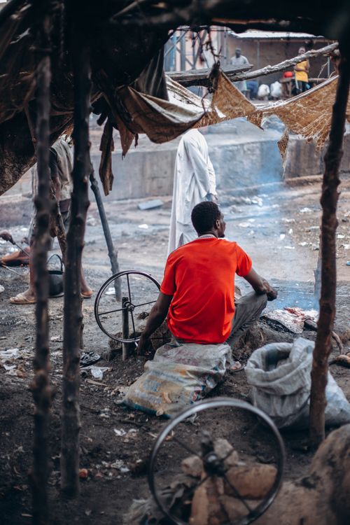 Free A man in a vibrant red shirt sits in a rustic urban environment, surrounded by makeshift structures. Stock Photo
