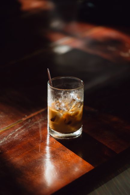 A glass of iced coffee with ice cubes sitting on a wooden table in warm sunlight.