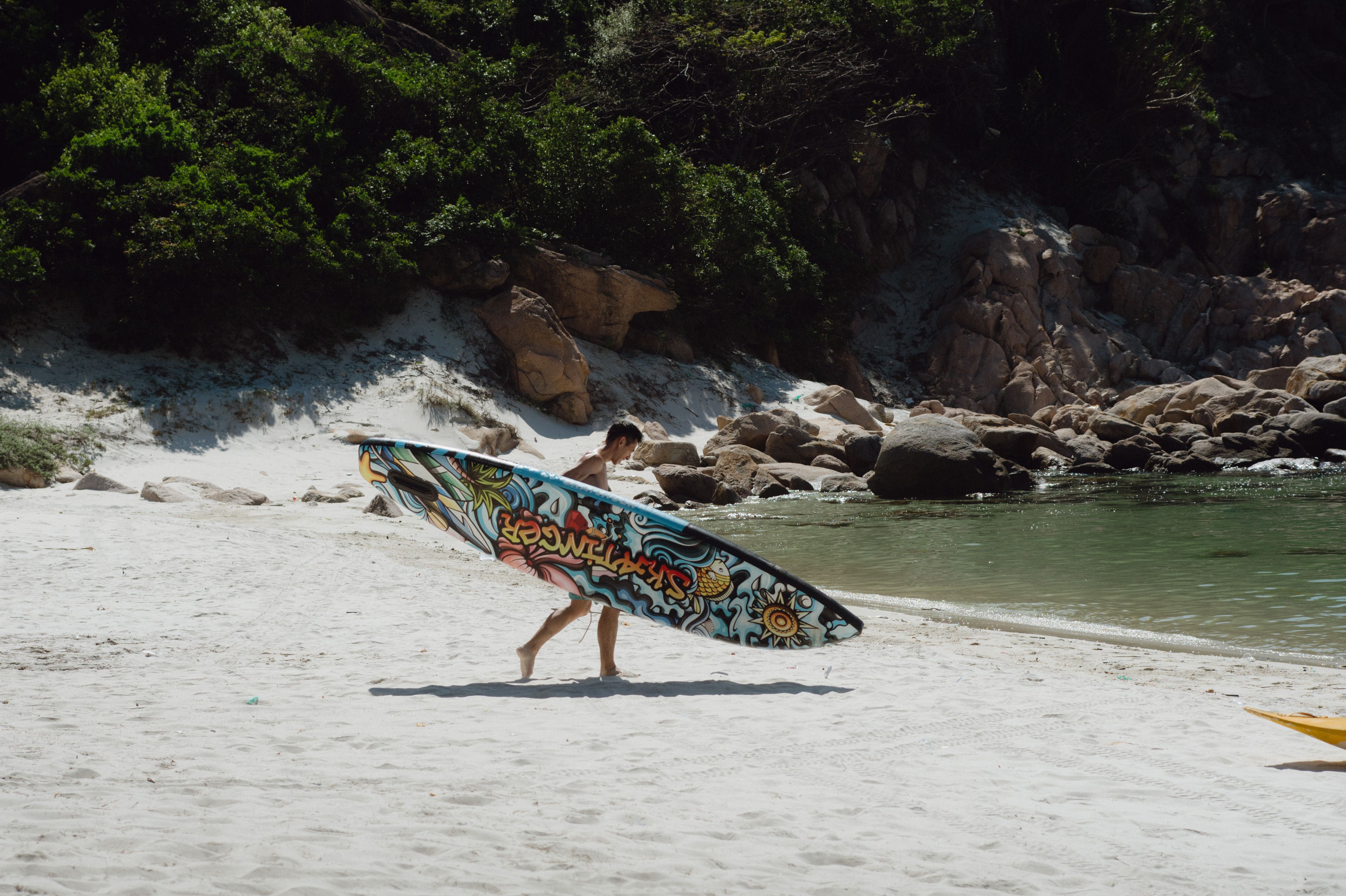 A man walking along a pristine beach carrying a vibrant, graffiti-style surfboard.