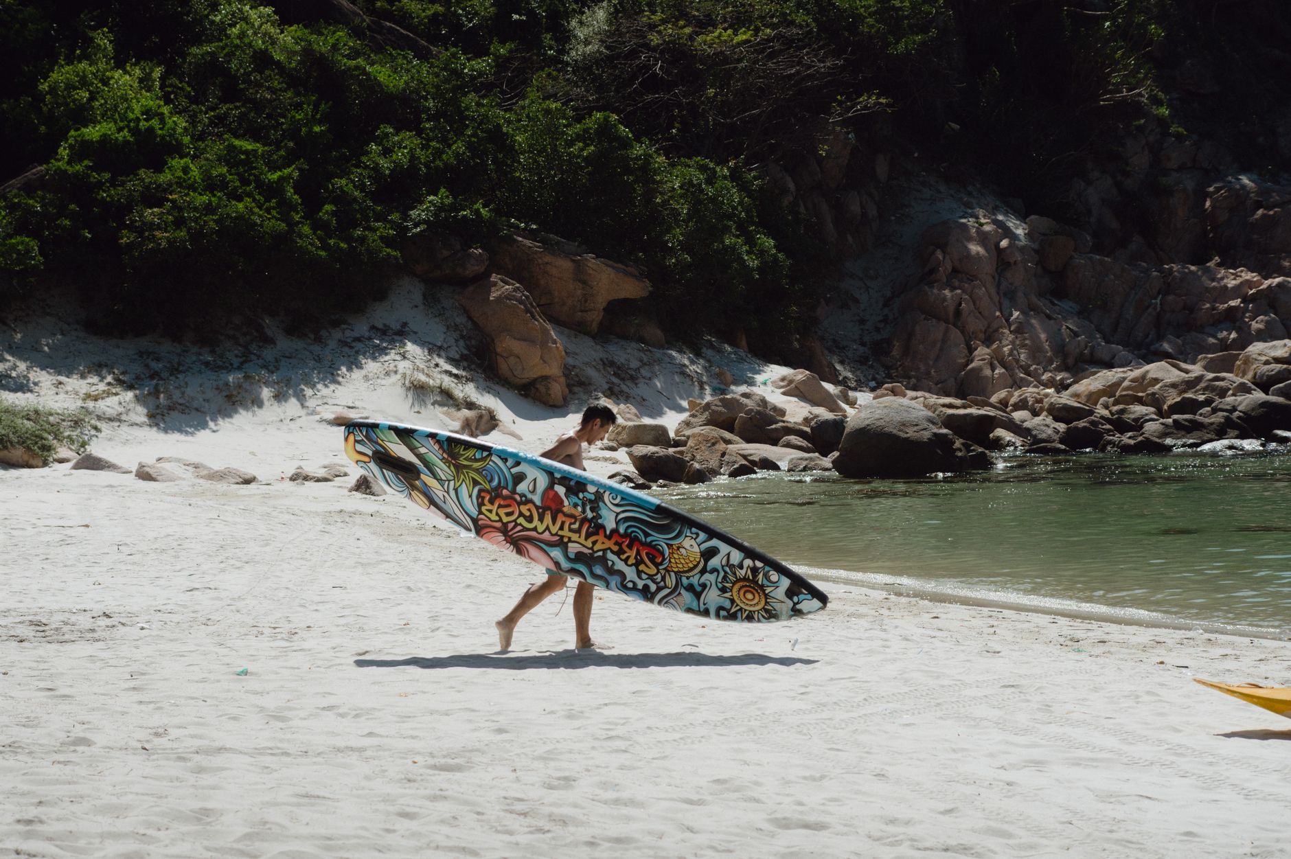 A man walking along a pristine beach carrying a vibrant, graffiti-style surfboard.