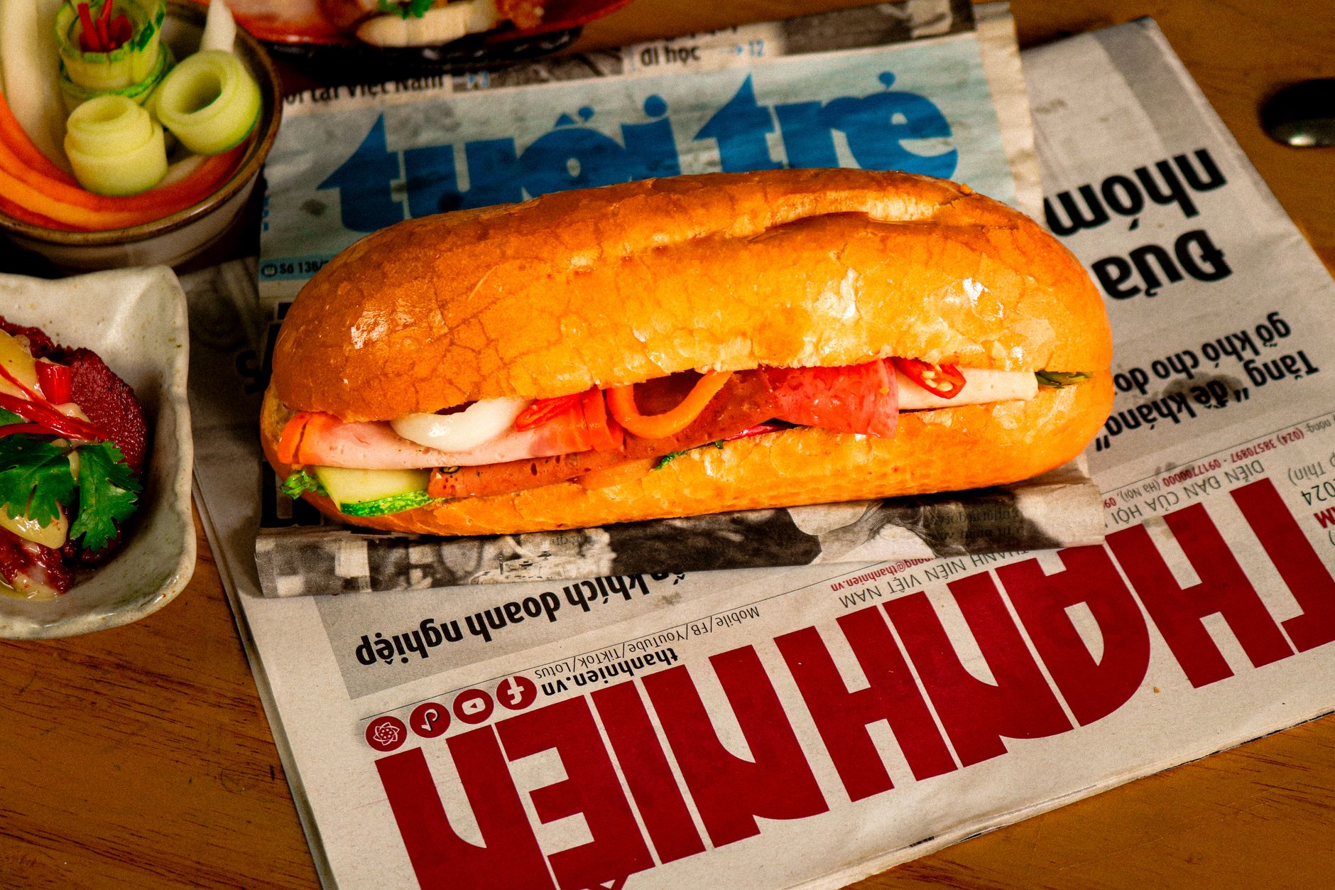 Close-up of a traditional Vietnamese bánh mì sandwich placed on a newspaper with side dishes.