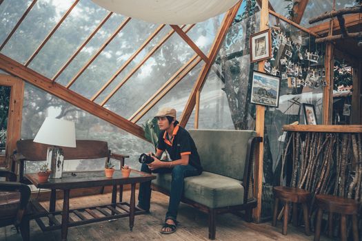 Young man holding a camera in a cozy, artistic indoor space with wooden furniture.