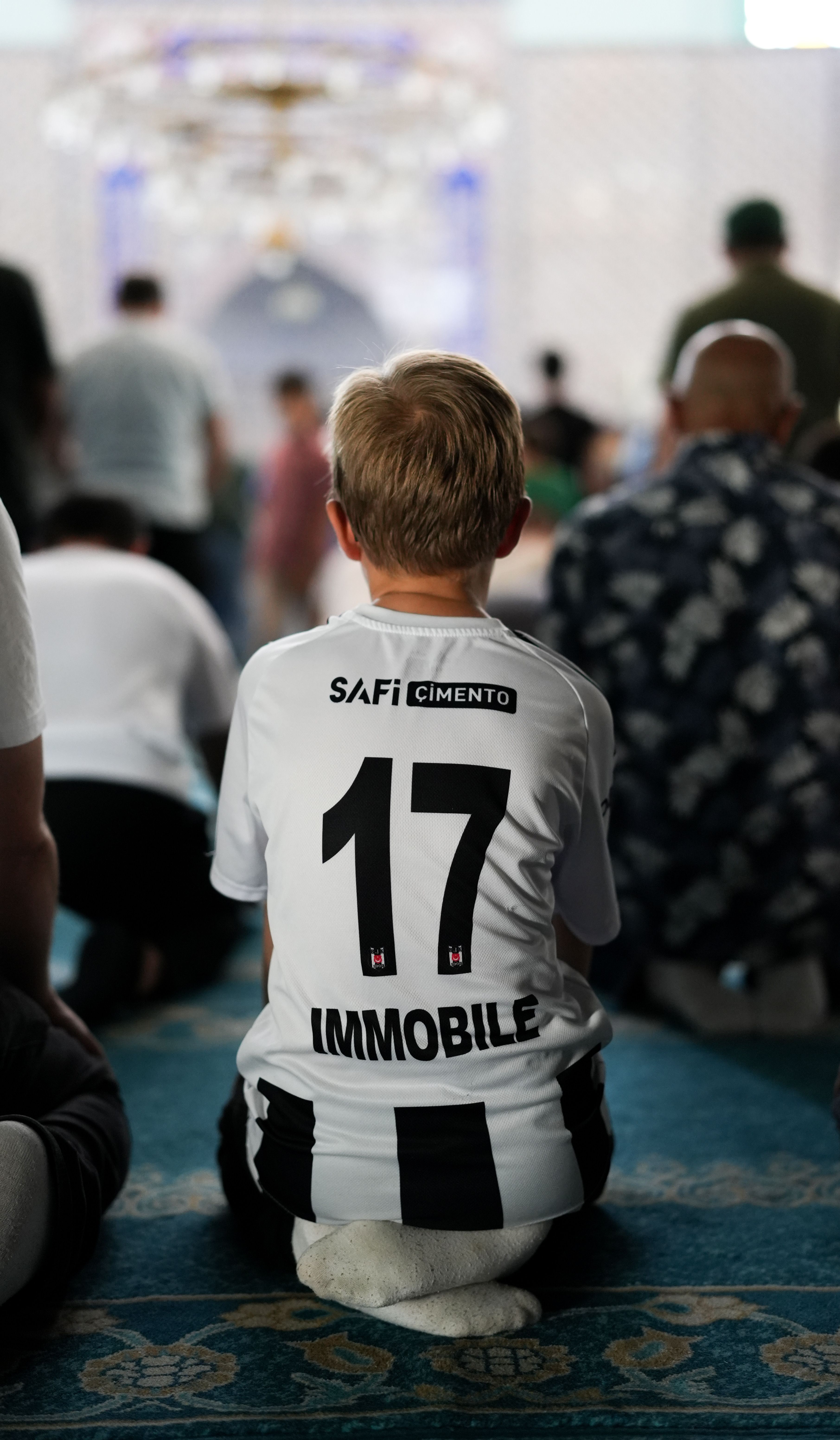 Child wearing a football jersey kneels in prayer at a mosque, blending sports and spirituality.