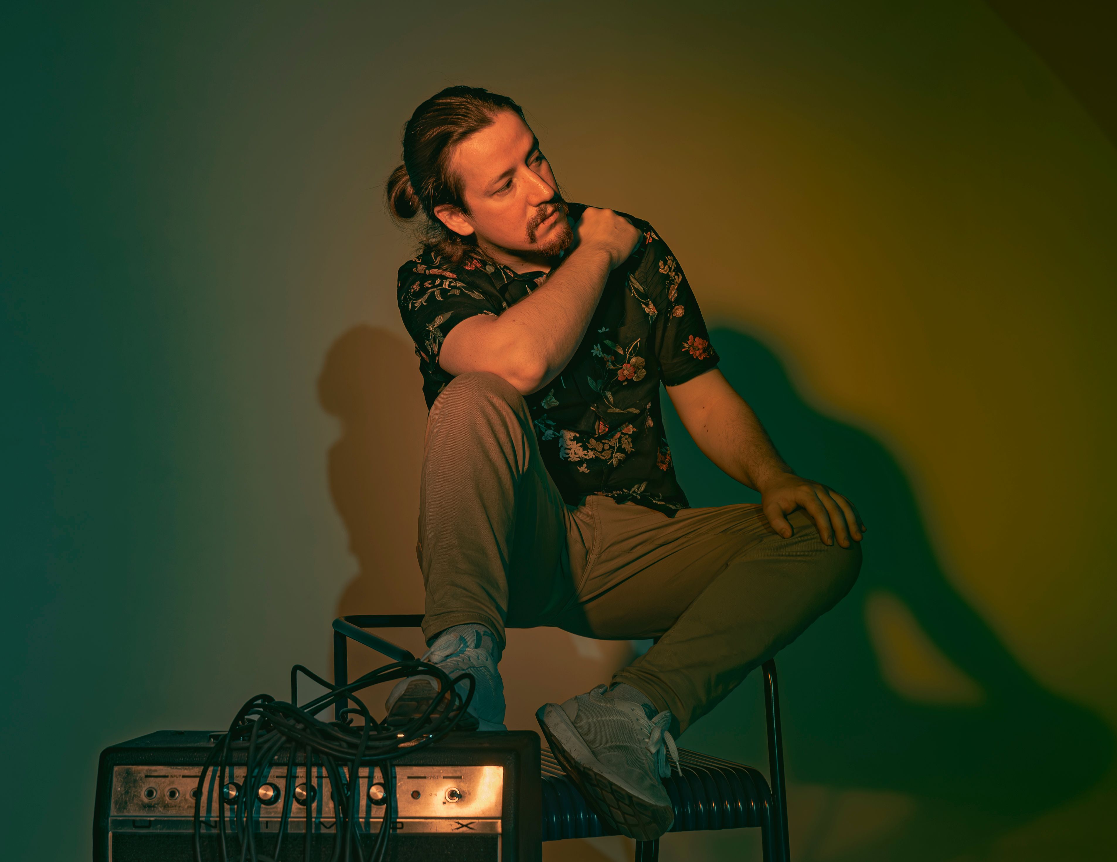 A thoughtful man sits by an amplifier, casting vivid shadows in warm lighting.