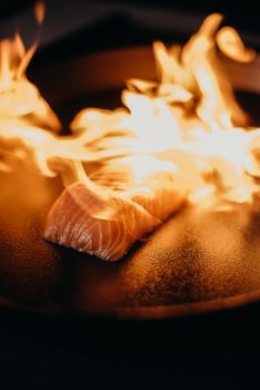 Close-up of a salmon fillet being seared with flames on a black iron pan, highlighting culinary artistry.