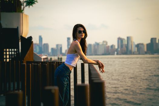A stylish woman in sunglasses leans against a railing with the New York City skyline in the background at sunset.