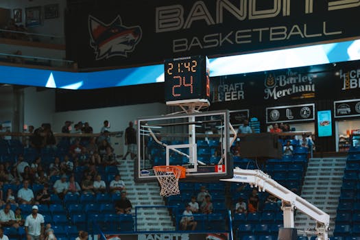 Close-up of a basketball hoop inside a stadium with a scoreboard and audience in the background.