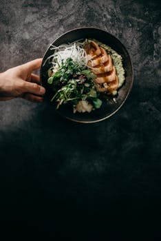 A hand presents a gourmet Asian fusion dish featuring grilled chicken and fresh greens.