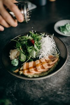 A tempting salmon dish garnished with greens, radish, and wasabi, being seasoned with sesame seeds.