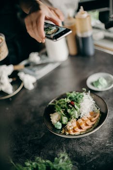 A person photographing a fresh salmon sushi dish garnished with greens and wasabi indoors.