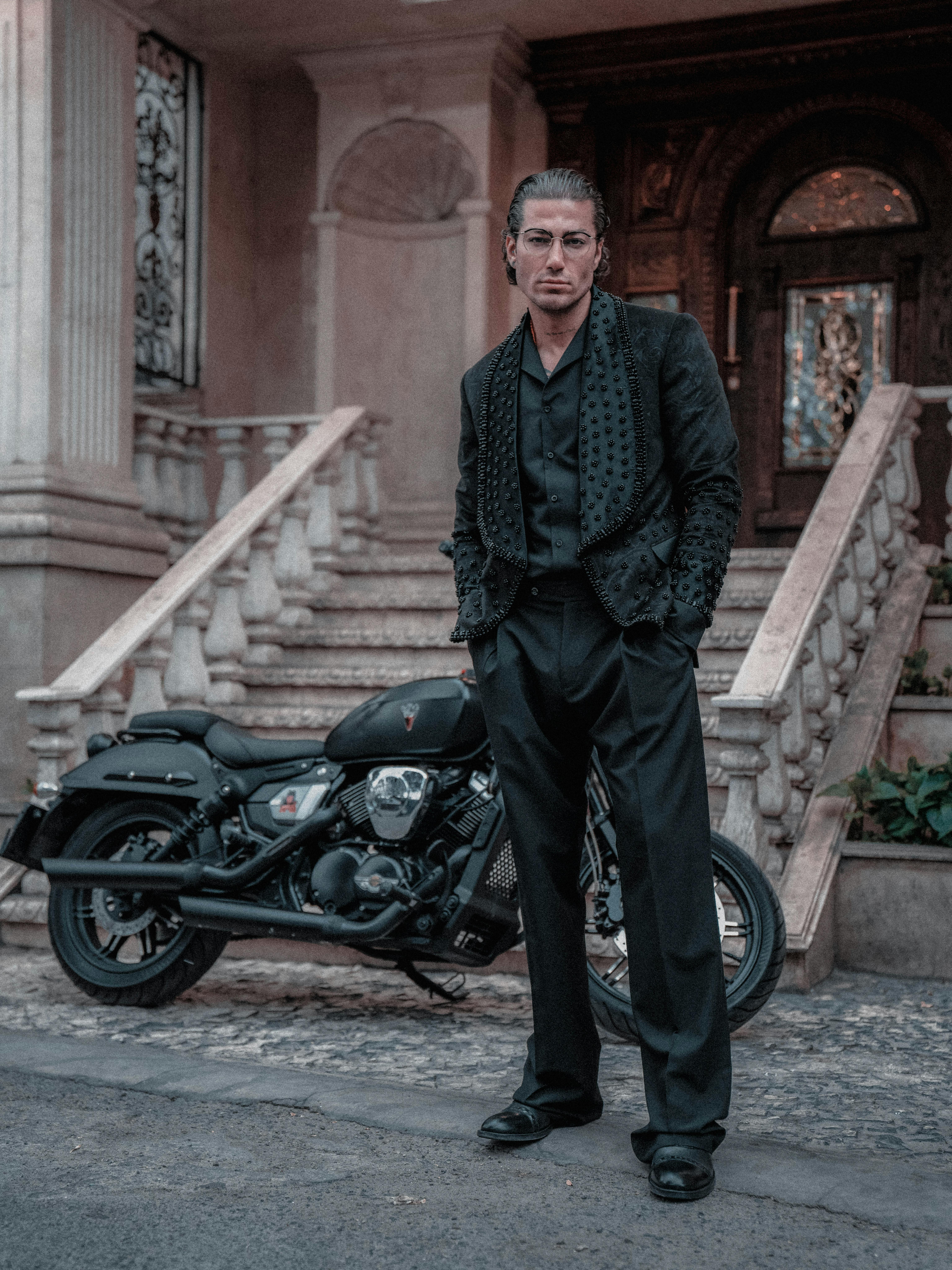 Stylish man in black attire standing confidently next to a black motorcycle on steps.