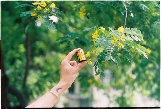 A hand with a moon tattoo holds a film roll against a backdrop of lush green leaves and yellow flowers.