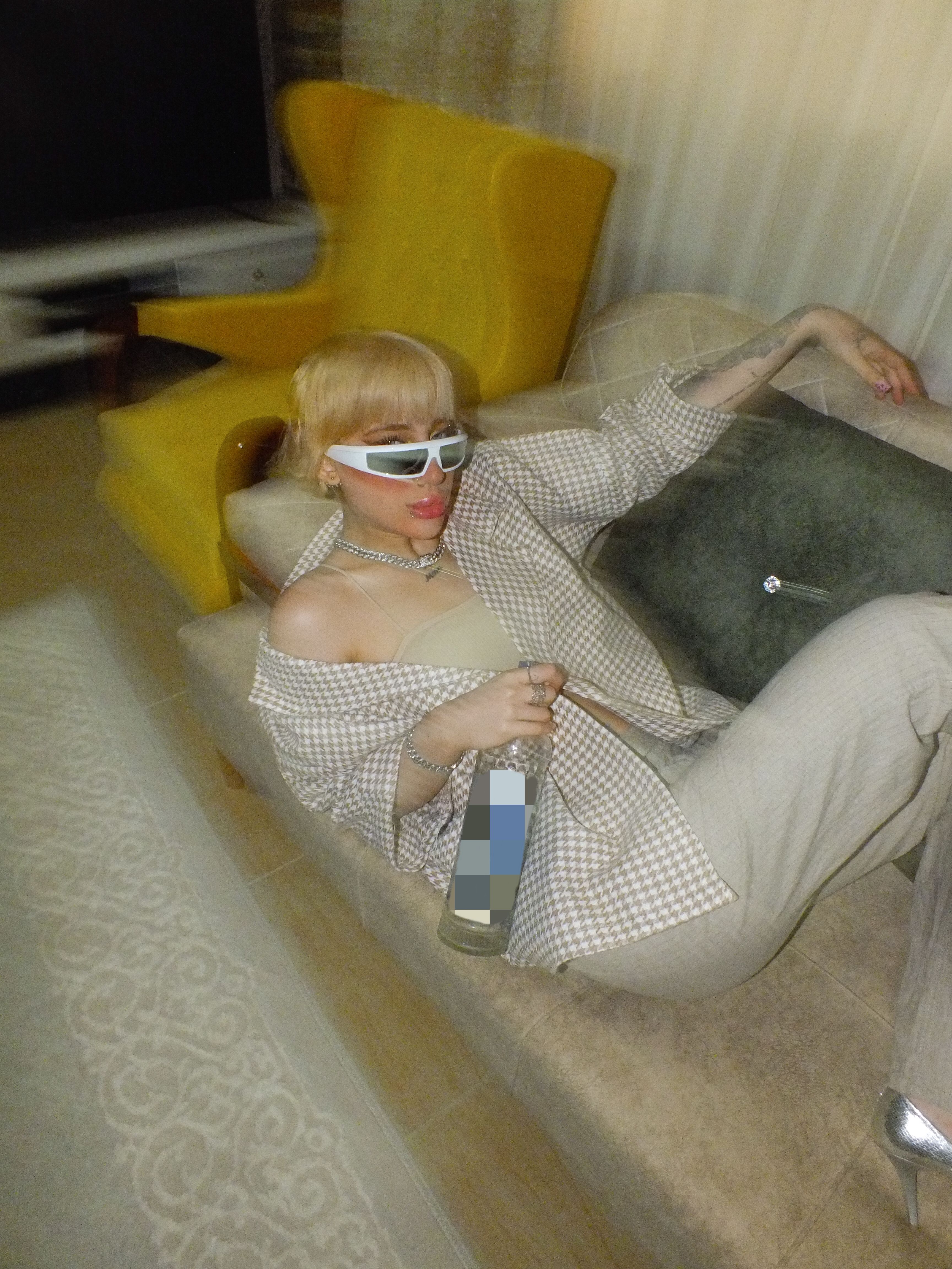 A stylish woman in sunglasses lounging indoors holding a drink, showcasing modern fashion and relaxation.