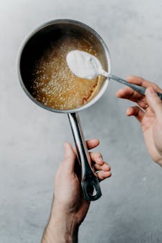 Close-up of hands cooking sugar mixture in saucepan indoors.