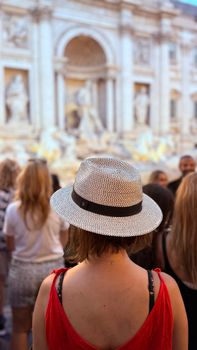 Back view of a woman in a straw hat admiring the iconic Trevi Fountain in Rome.