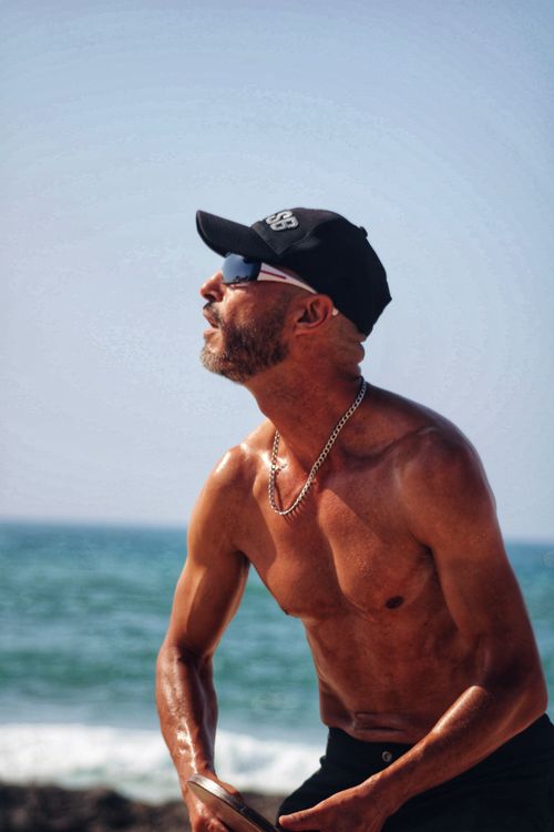 Free Sunlit portrait of a muscular man on the beach, wearing sunglasses and cap, enjoying a summer day. Stock Photo