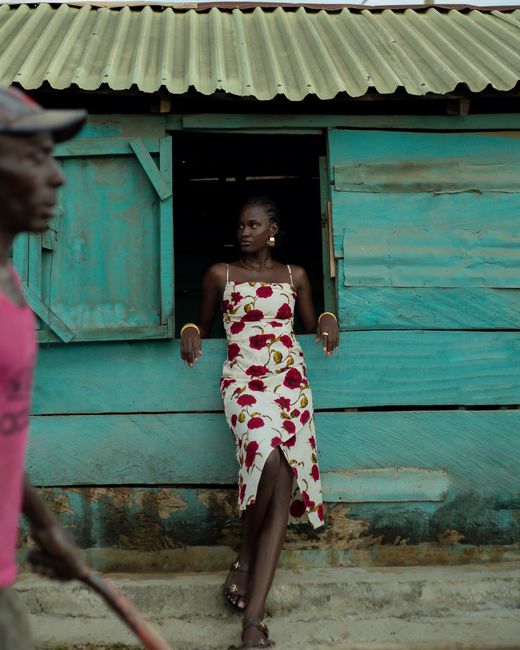 A stylish woman in a floral dress leans against a rustic teal wall in Ilorin, Nigeria.