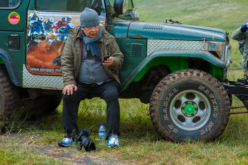 Free Man sitting by a jeep with a camera, checking phone amidst grassy outdoors. Stock Photo