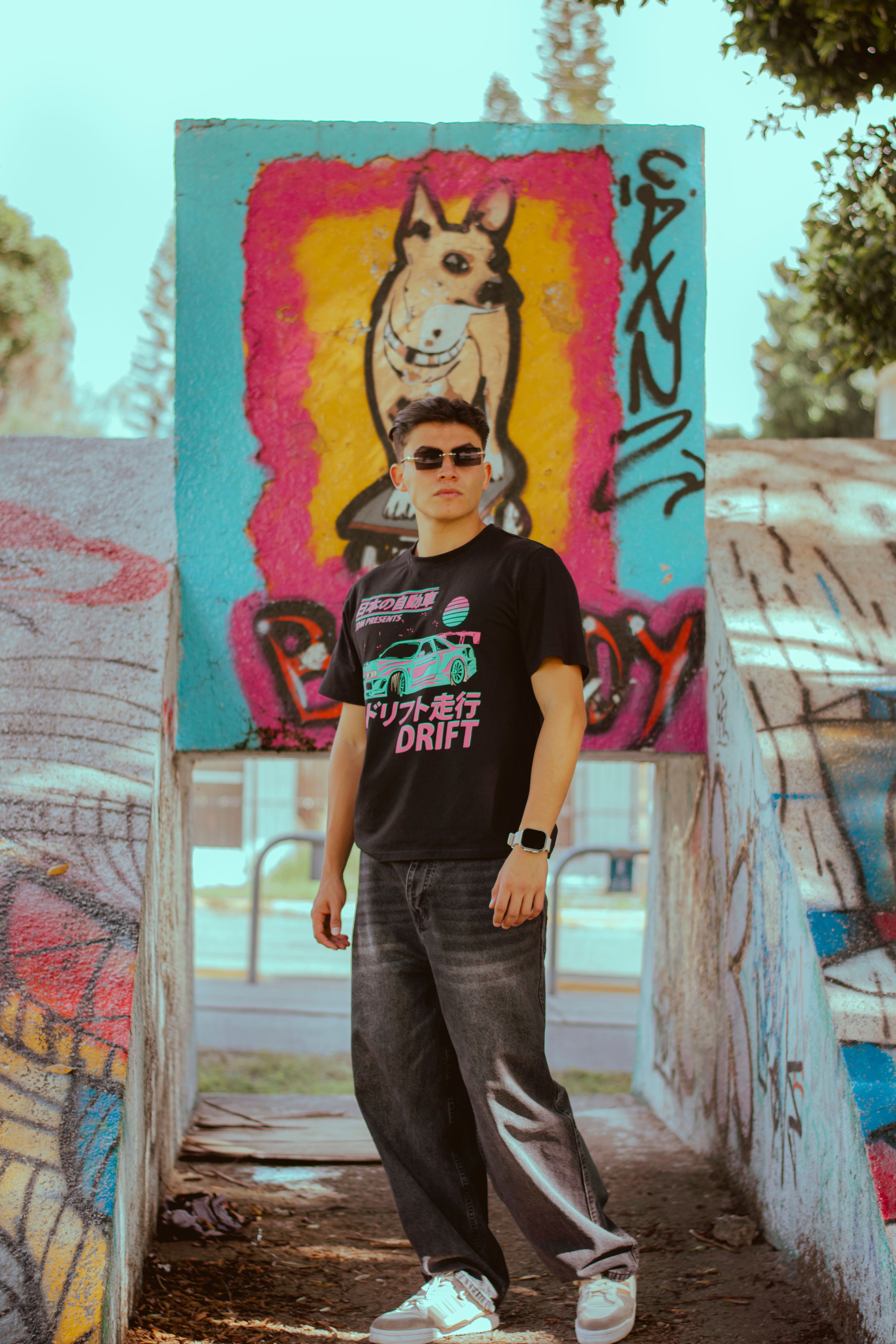 A stylish young adult poses in front of a vivid graffiti mural outdoors during the day.