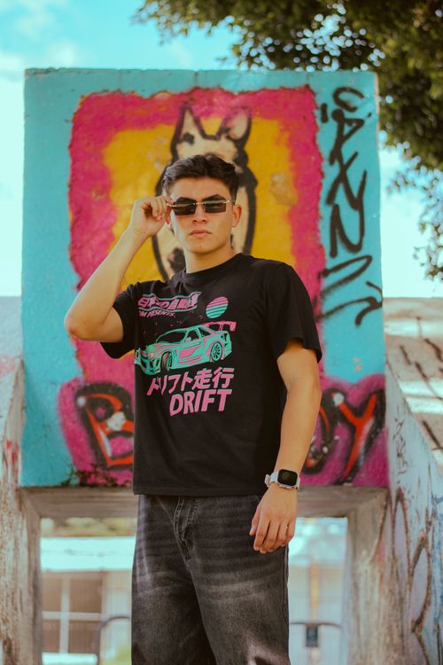 Free Confident young adult wearing sunglasses and graphic tee in front of vibrant graffiti wall. Stock Photo