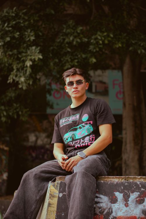 Free Stylish young man wearing sunglasses and casual wear sitting by graffiti wall in urban setting. Stock Photo