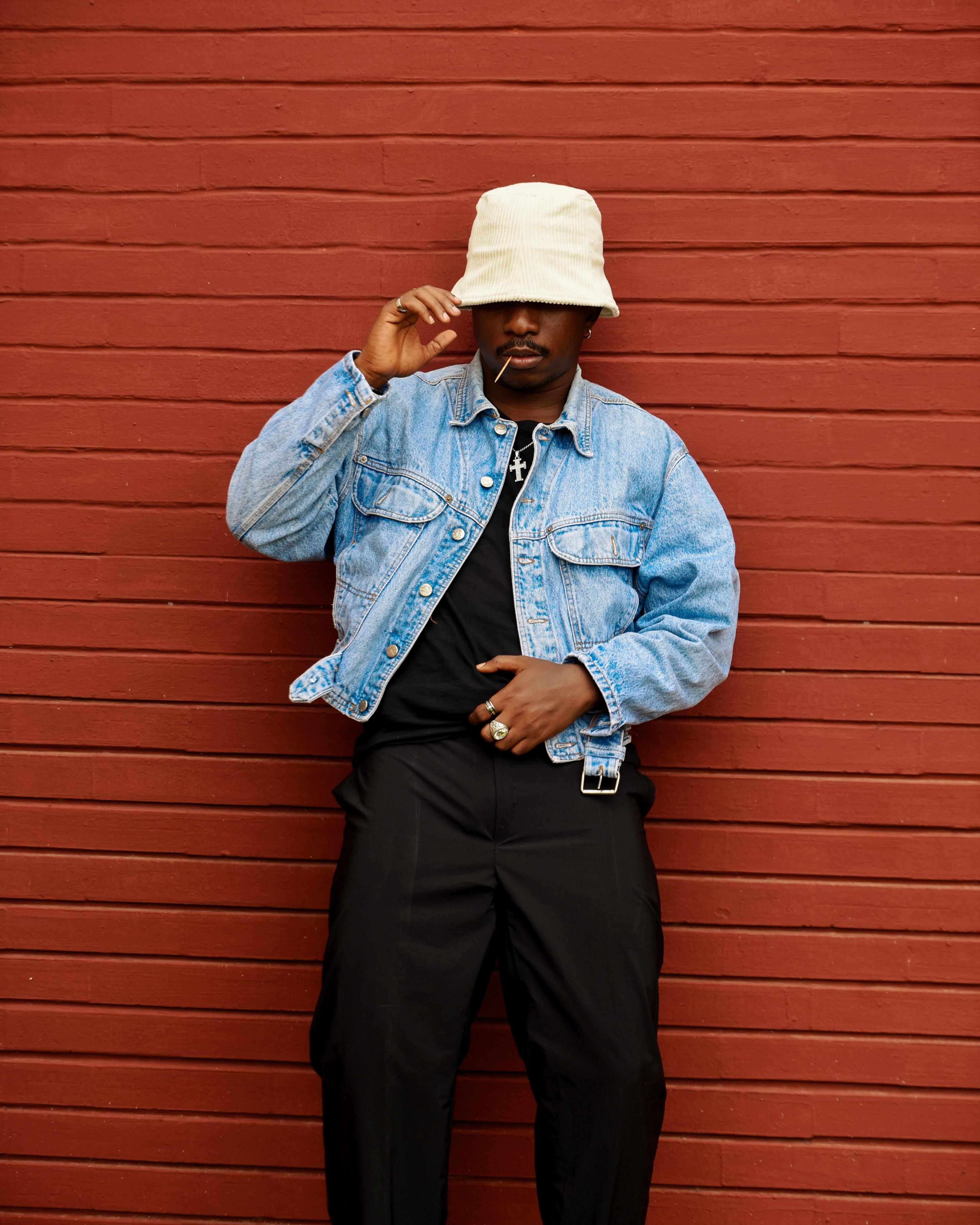 Man with denim jacket and hat poses stylishly against a red brick wall.