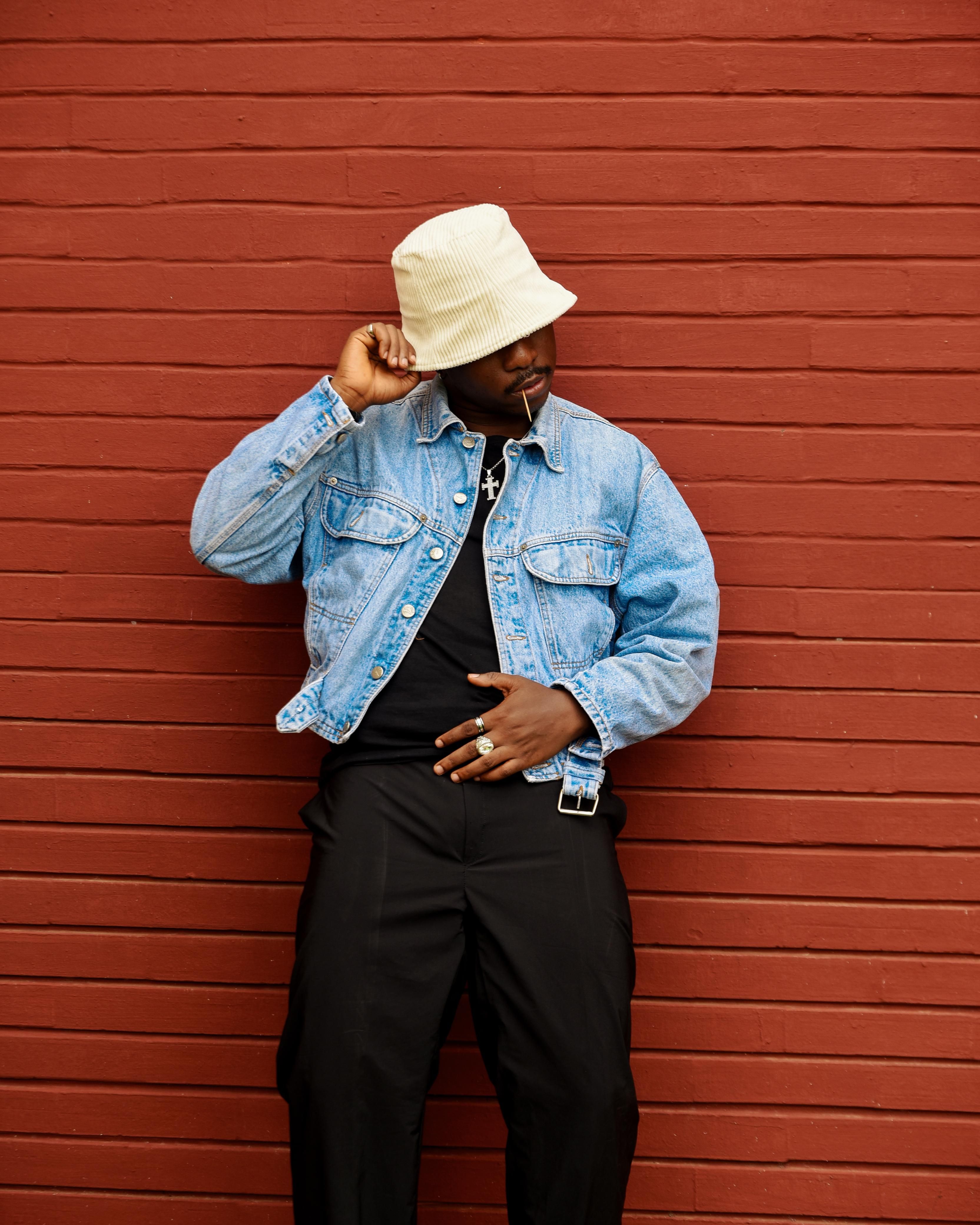 Stylish man wearing a denim jacket and hat posing confidently against a red brick wall.