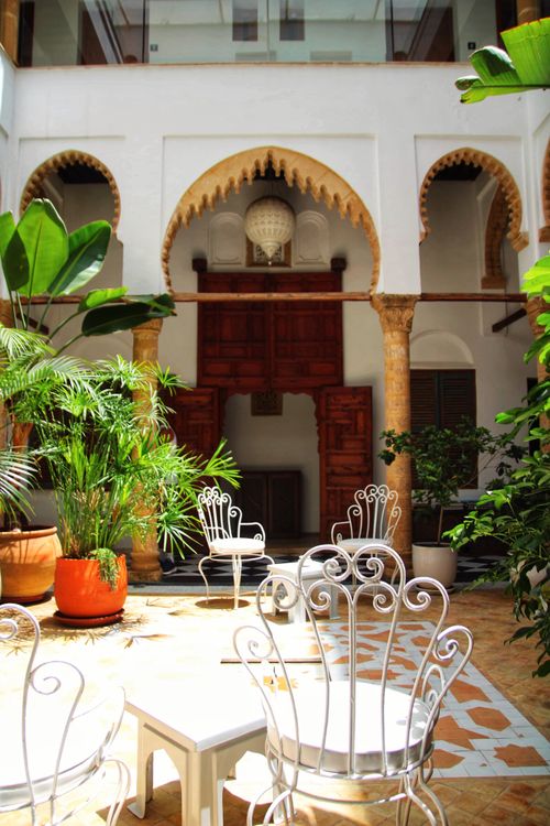 Free Serene Mediterranean courtyard featuring ornate arches, lush plants, and elegant white chairs under bright daylight. Stock Photo