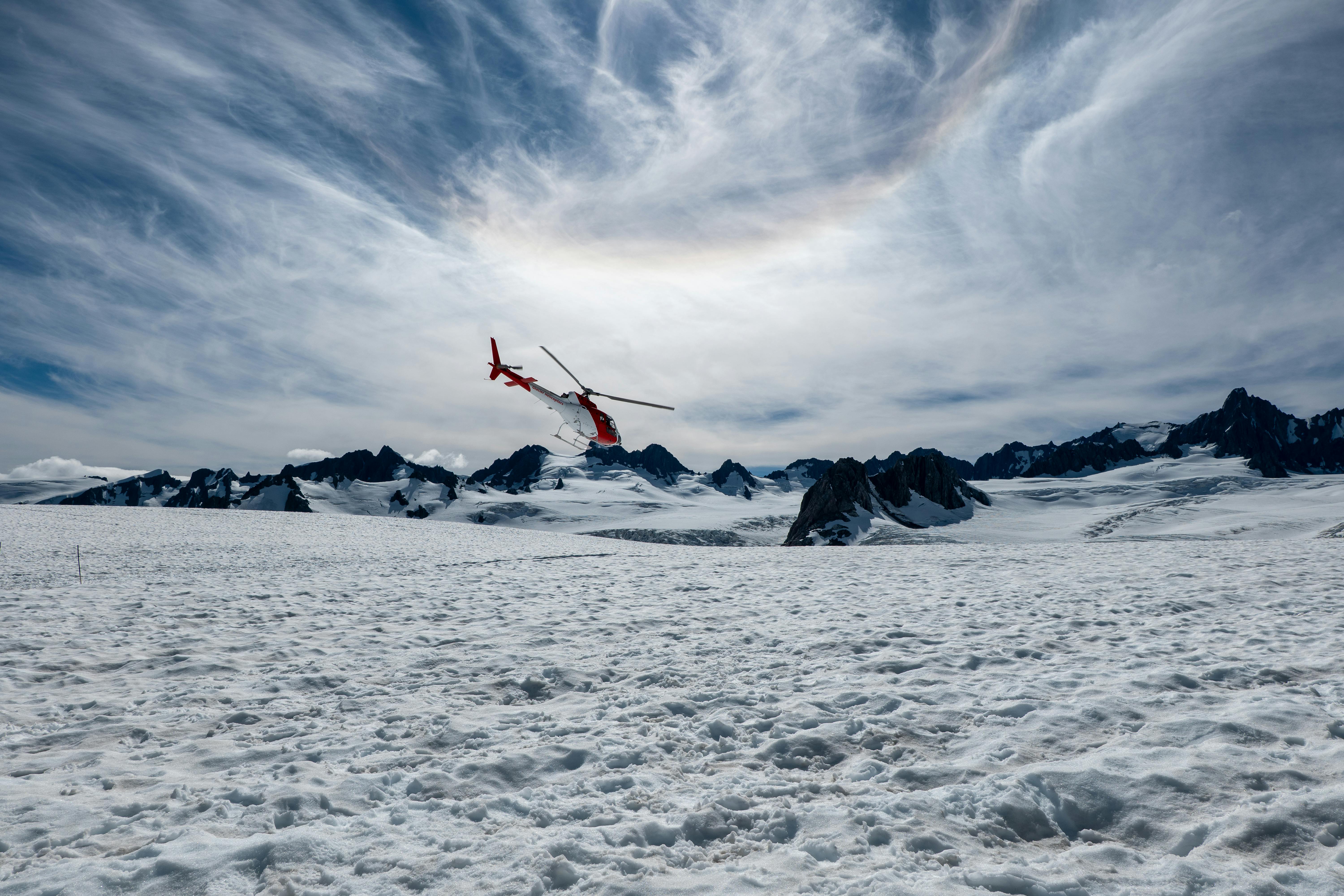 A helicopter hovers over a stunning snowy landscape in New Zealand with dramatic skies.