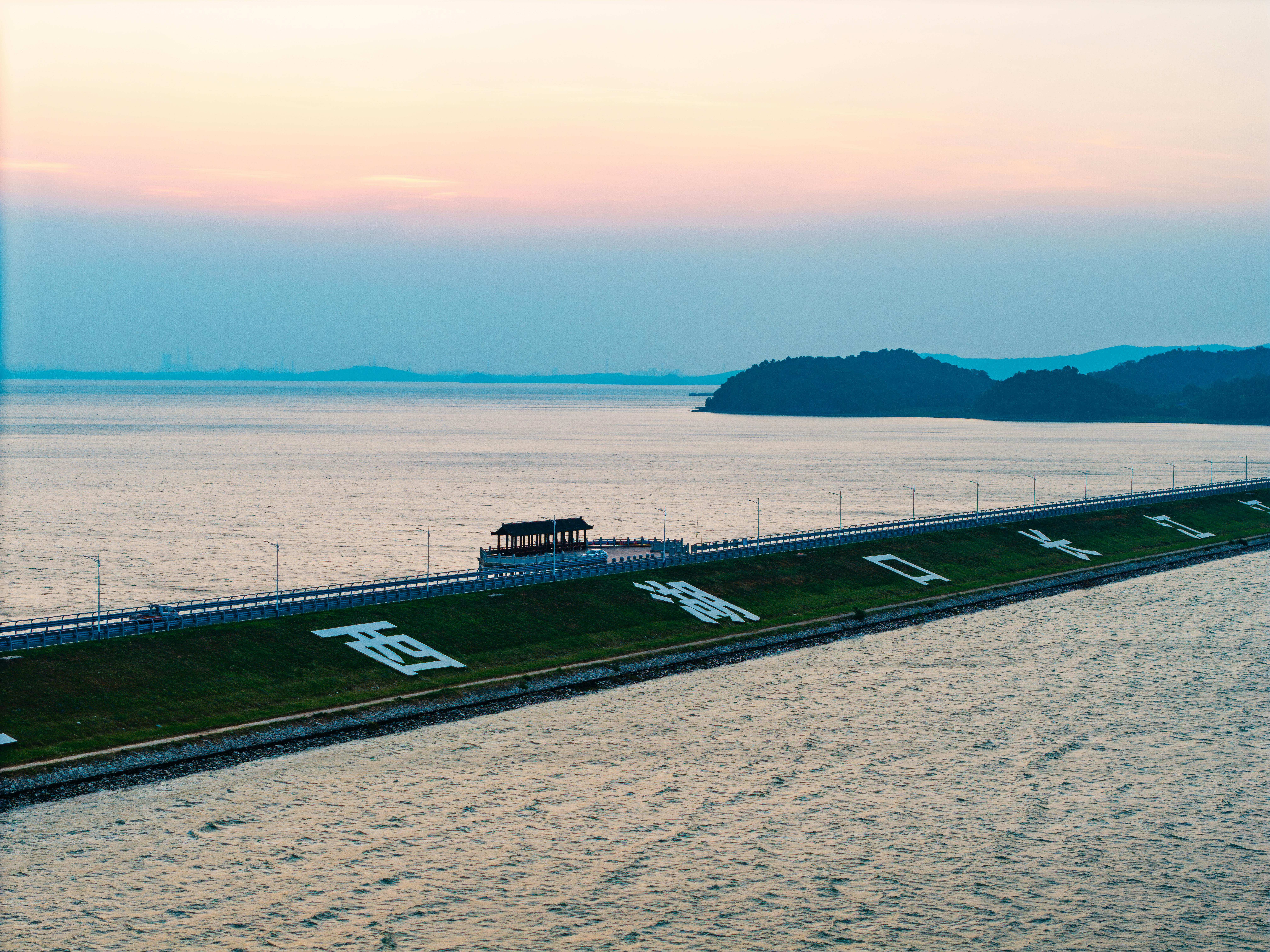 A serene lakeside scene with a dam, pavilion, and hilly backdrop during sunset.