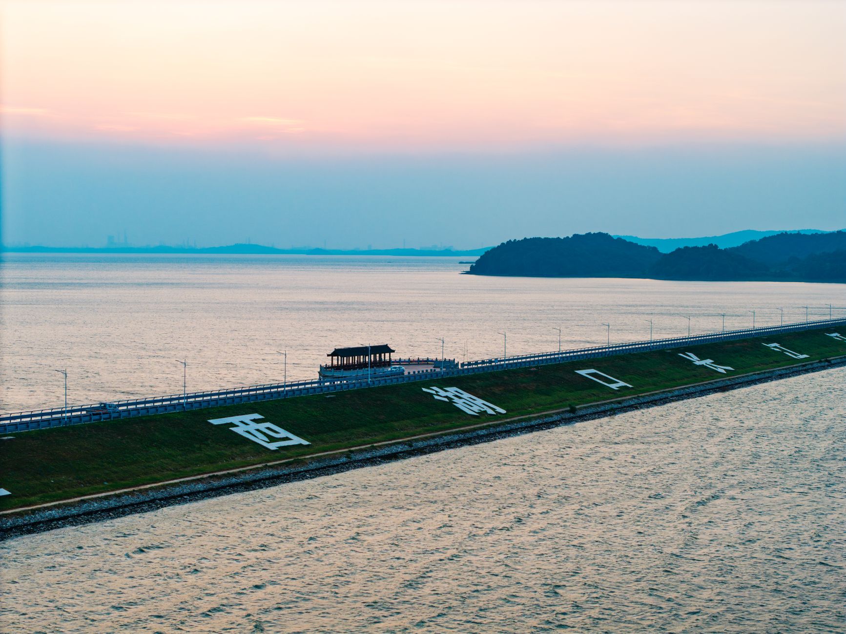 A serene lakeside scene with a dam, pavilion, and hilly backdrop during sunset.
