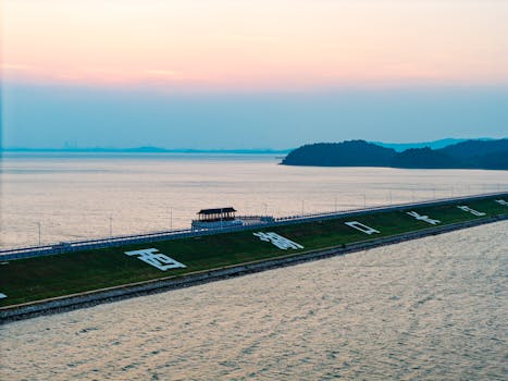 A serene lakeside scene with a dam, pavilion, and hilly backdrop during sunset.