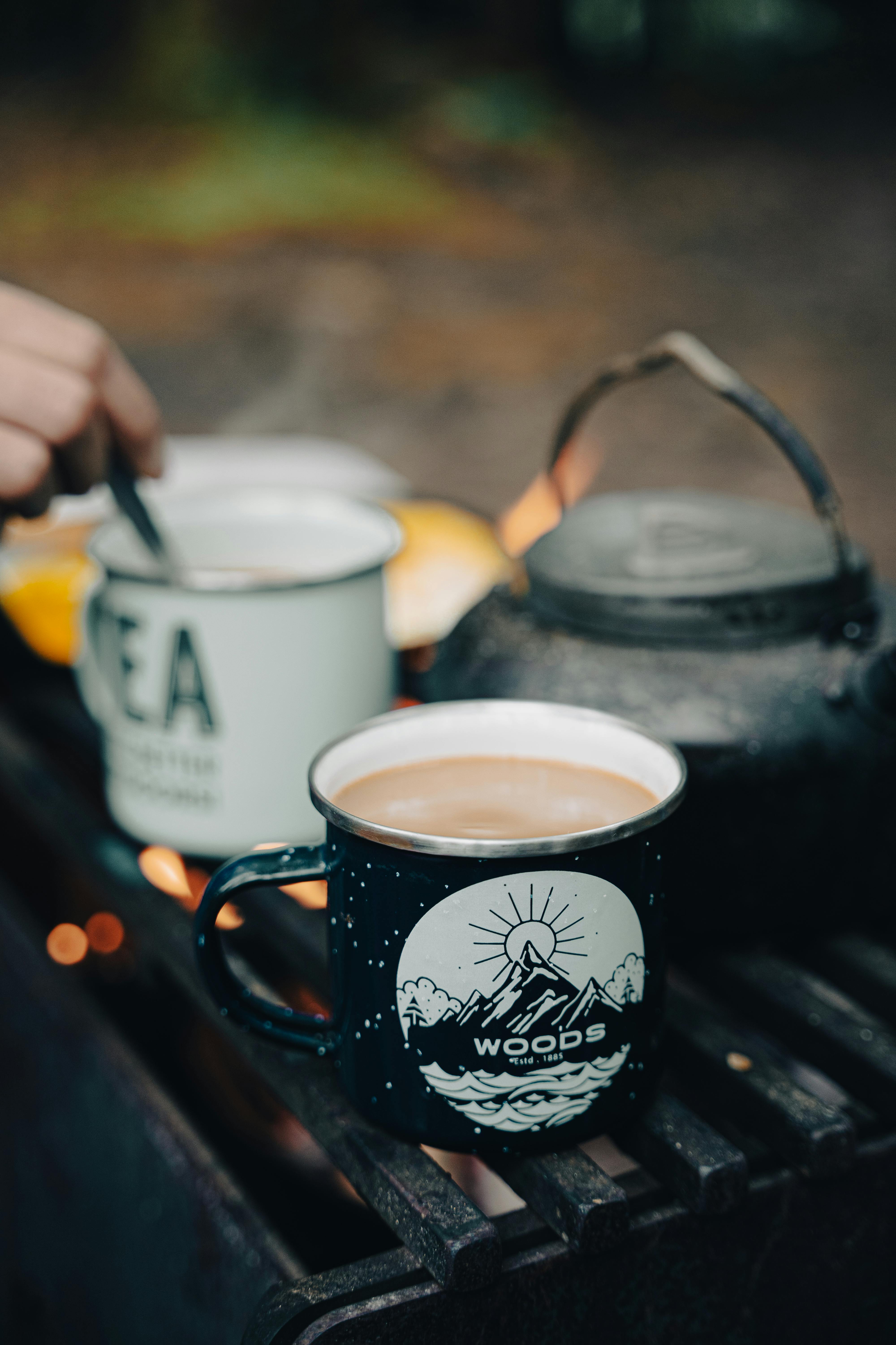 Close-up of tea and coffee mugs by a campfire, perfect for outdoor camping vibes.