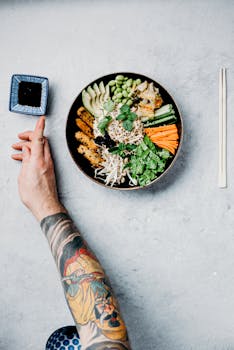 A vibrant Buddha bowl with fresh vegetables and a tattooed arm reaching for soy sauce.