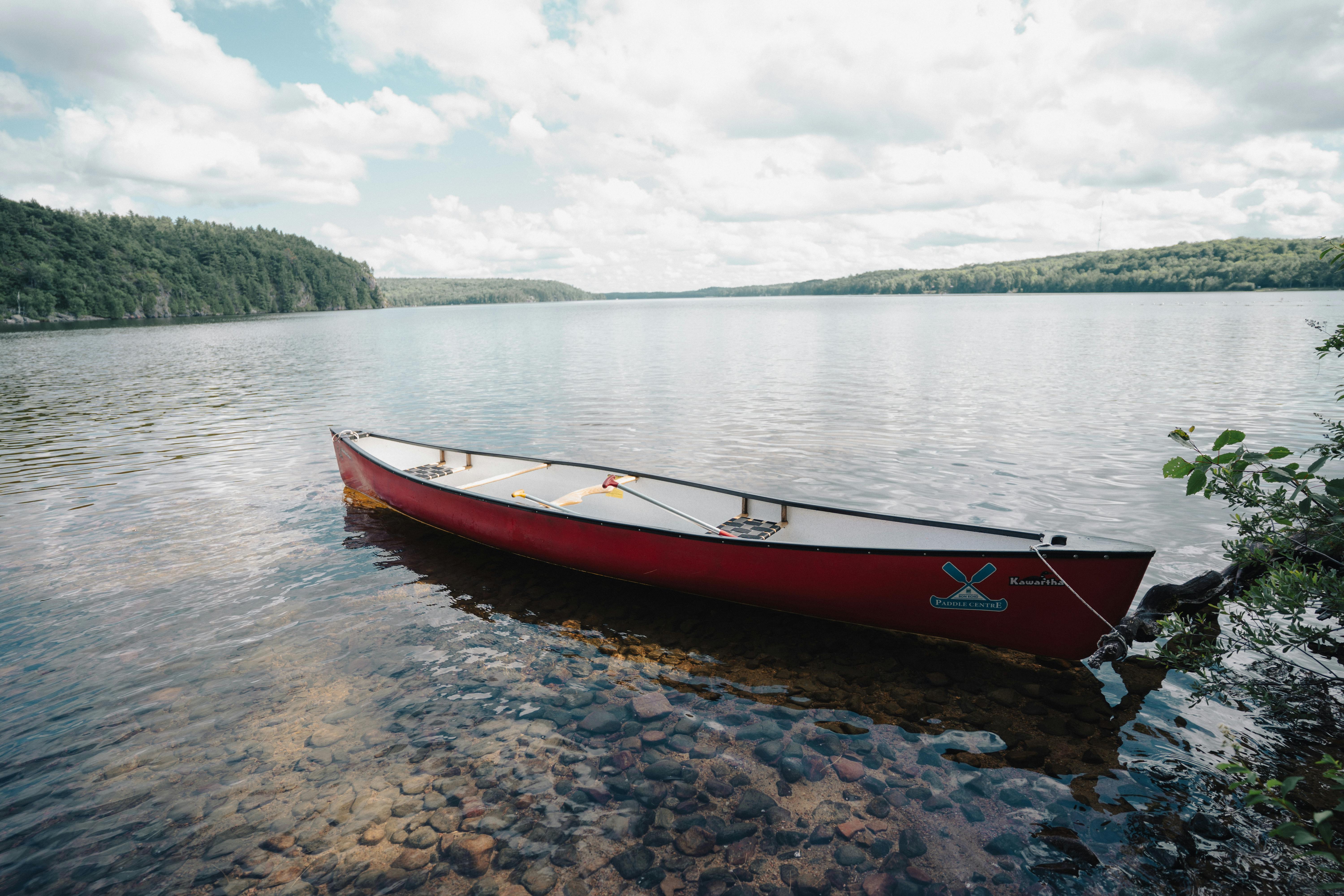 A serene red canoe rests on a calm lake's edge surrounded by lush, green forests under a cloudy sky.