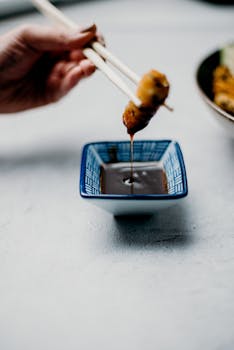 Close-up of a hand using chopsticks to dip food into a sauce bowl, showcasing Asian cuisine.