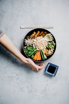 Top view of a colorful Asian bowl with fresh vegetables, edamame, sprouts, and soy sauce.