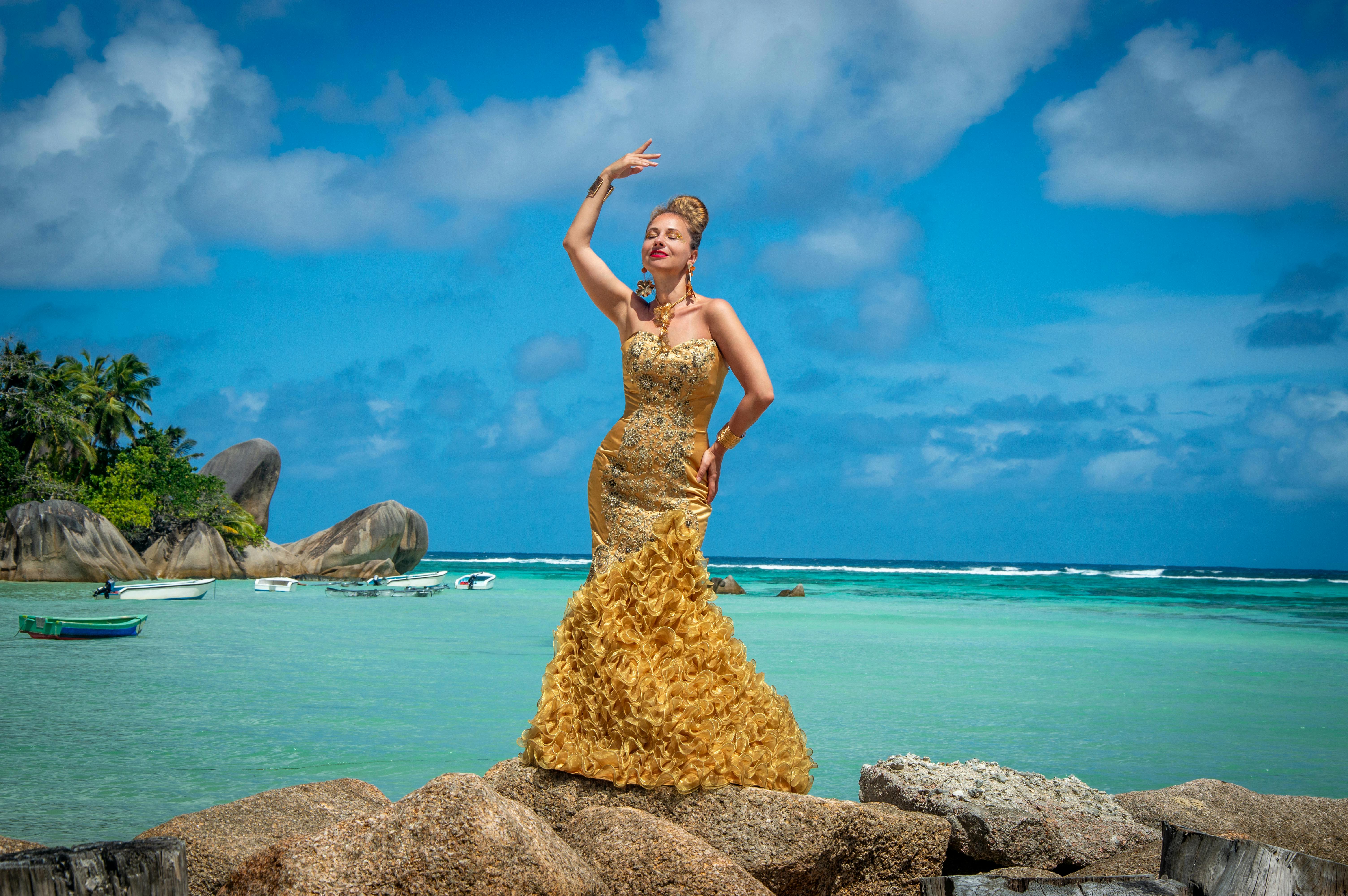 Woman in a luxurious gold dress posing on rocks by a tropical beach with clear blue sky.