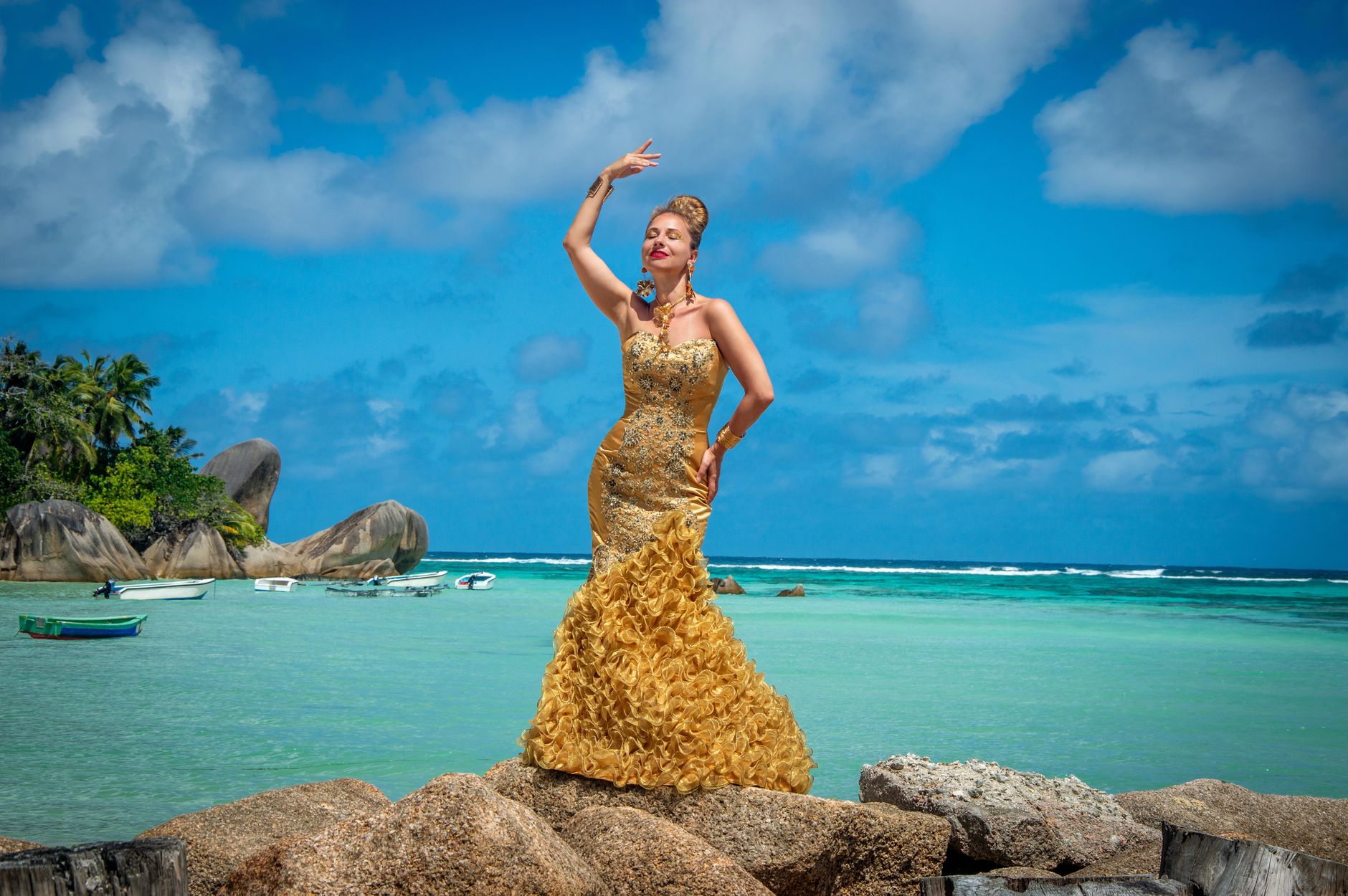 Woman in a luxurious gold dress posing on rocks by a tropical beach with clear blue sky.