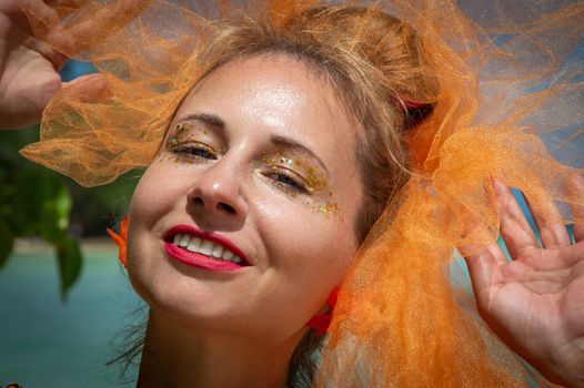 Close-up of a woman smiling with glitter makeup and an orange tulle veil, enjoying a sunny day.