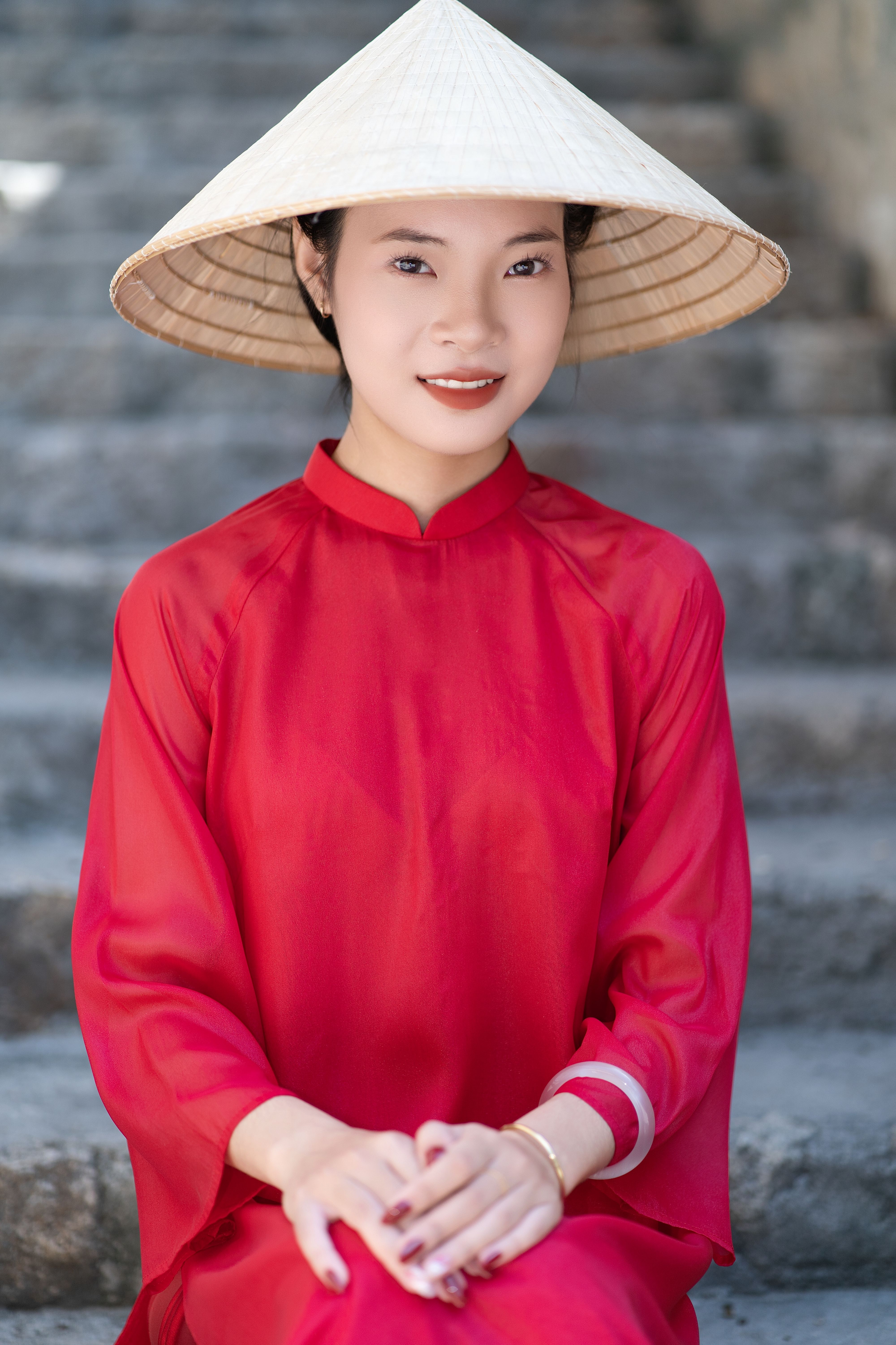Graceful woman in red ao dai and conical hat sitting on stone steps outdoors.