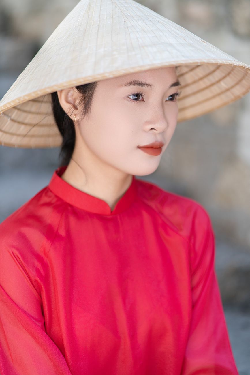 Elegant portrait of a woman in a red áo dài and conical hat.