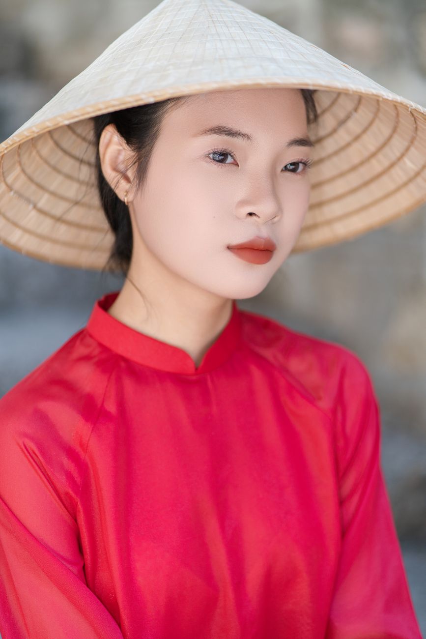 A close-up portrait of a woman wearing a traditional Vietnamese conical hat and red attire.