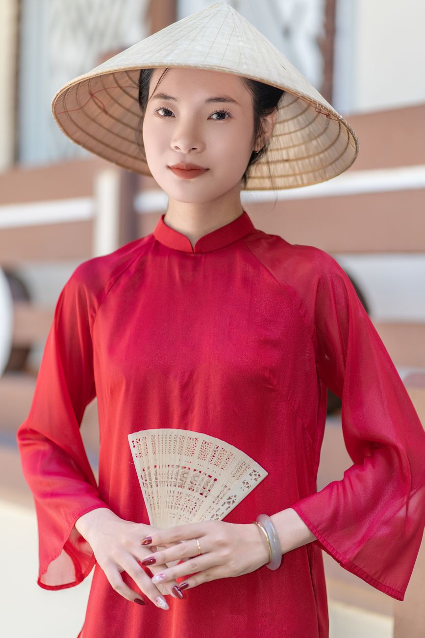 Portrait of an Asian woman in a traditional red dress and hat holding a decorative fan.